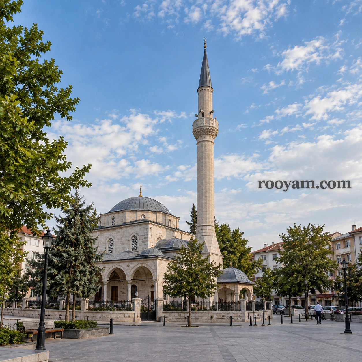Rüyada Minare: uzun taş bir minare, ağaçlar ve binalarla çevrili kubbeli bir caminin yanında yükseliyor.