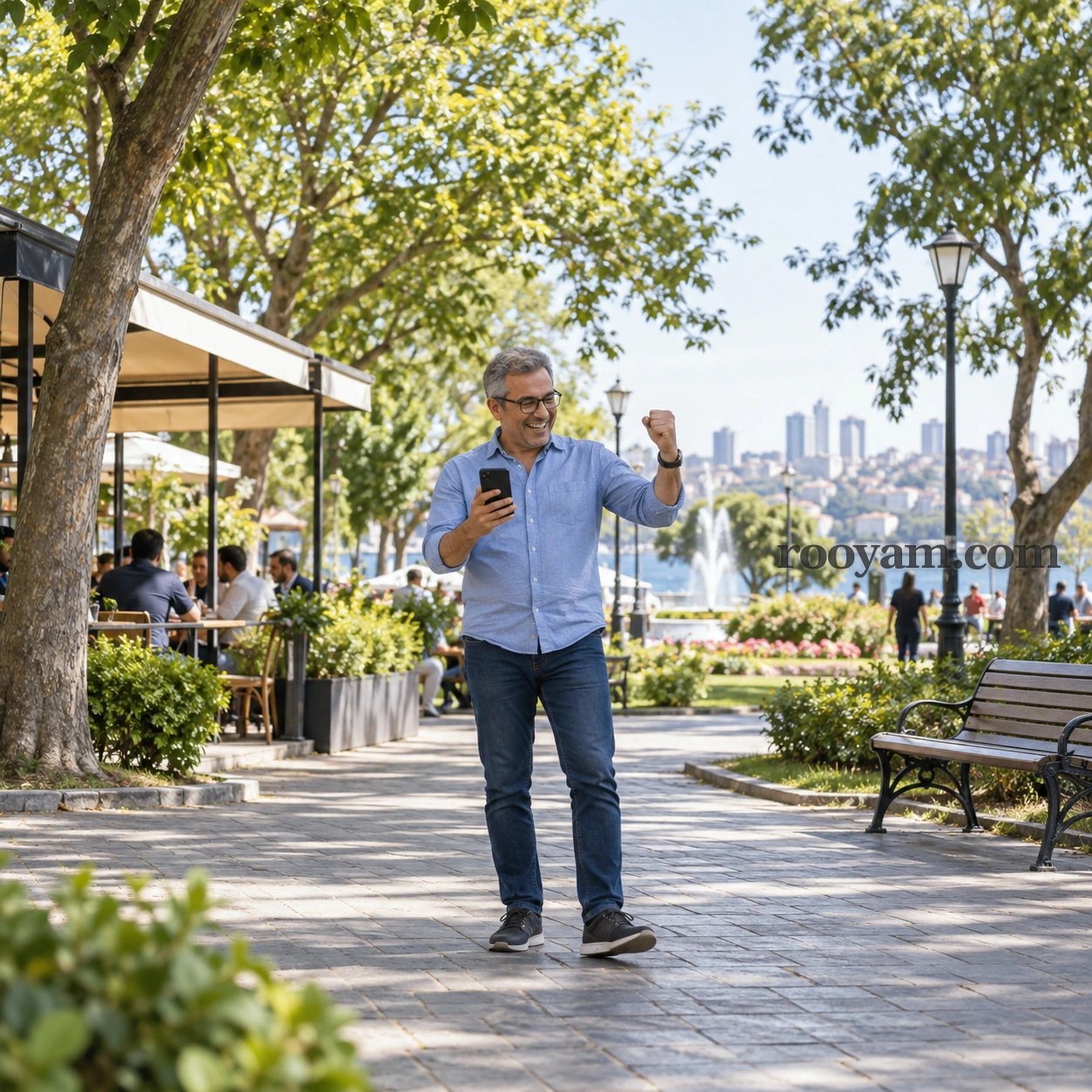 Rüyada Sevinmek: gülümseyen bir adam, ağaçlıklı park yolunda telefonunu tutup yumruğunu havaya kaldırıyor.
